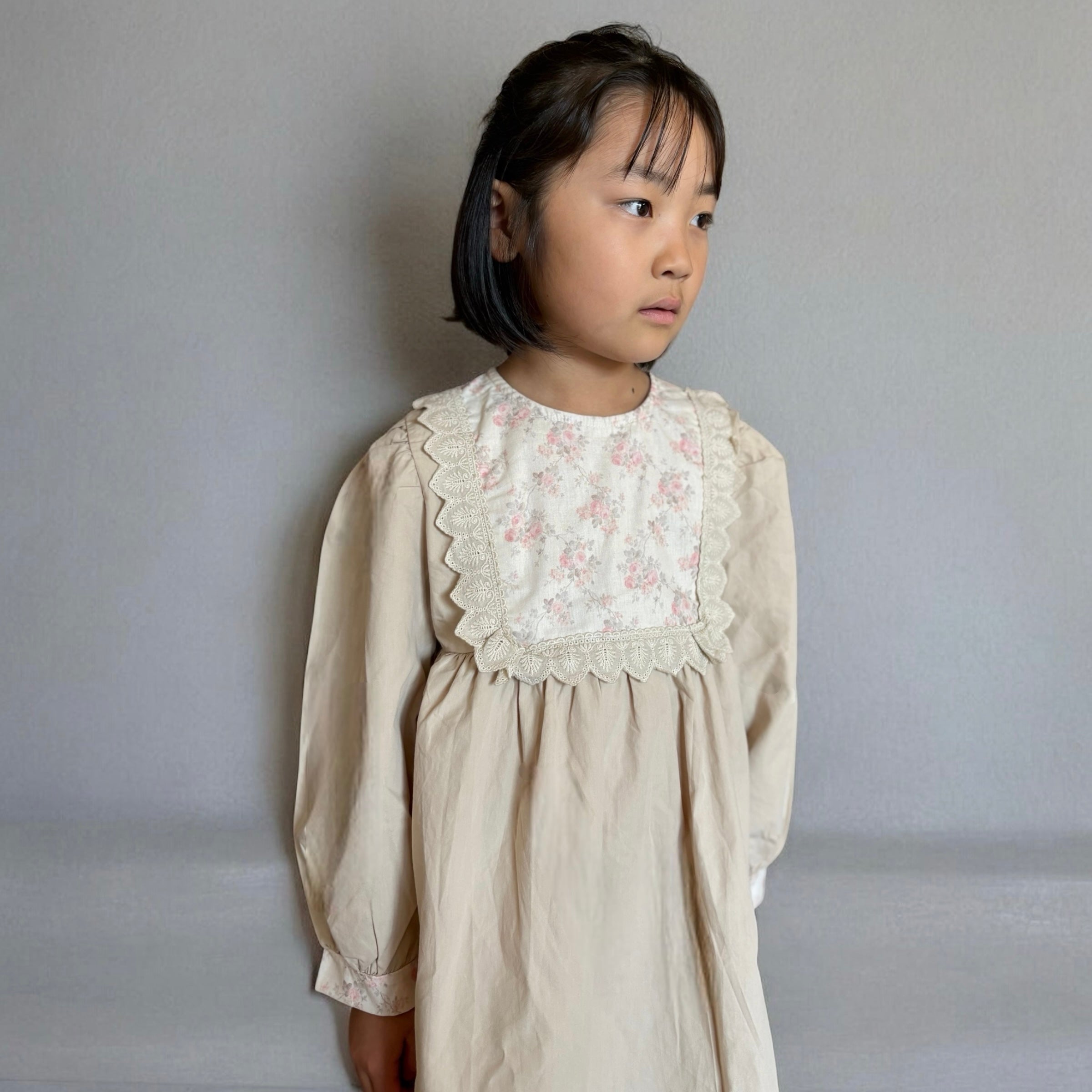 ベージュのクラシカルワンピースを着る女の子。胸元に小花柄とレースが施され、秋冬のお出かけや家族写真にぴったりの子供服